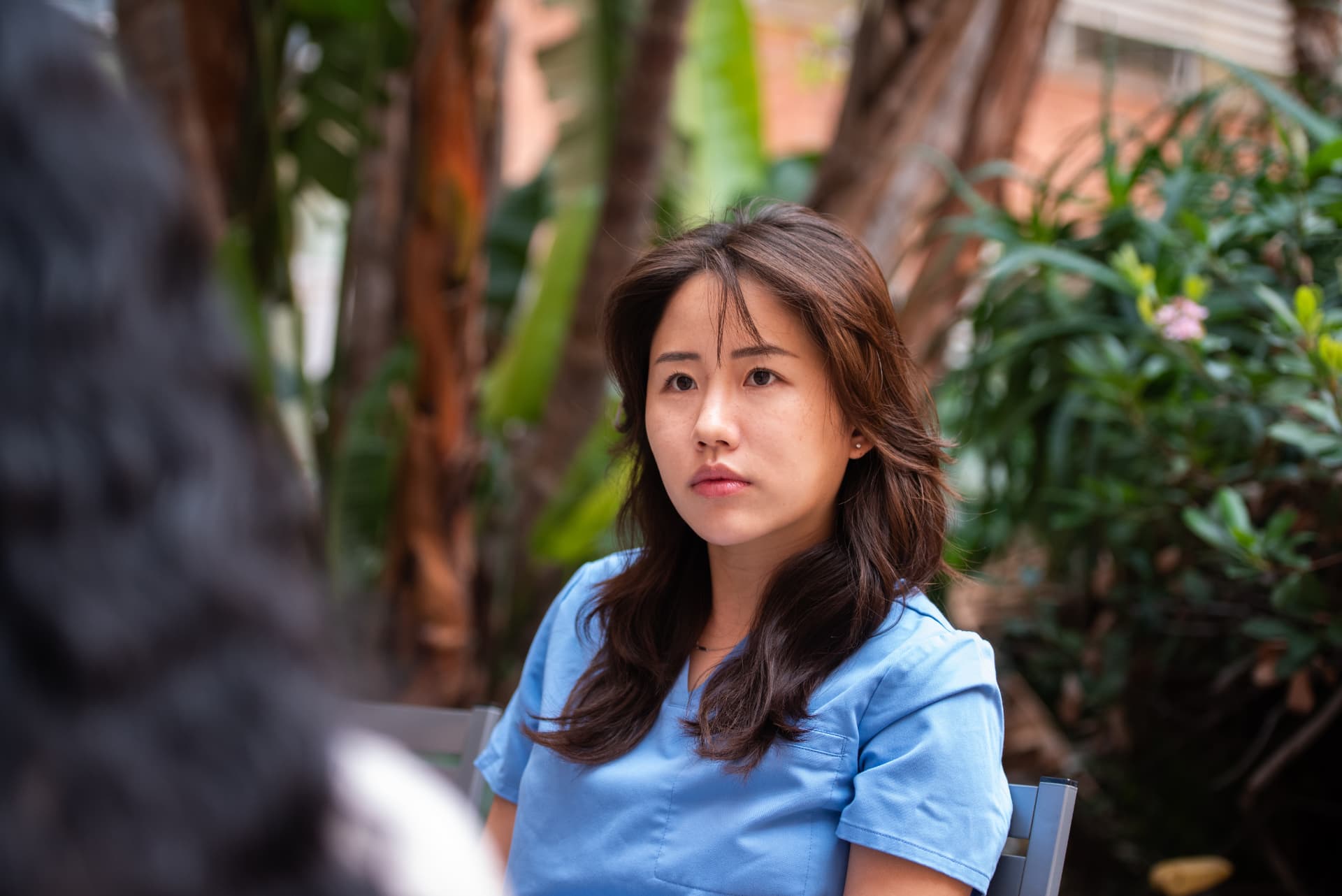 Nurse in blue scrubs seated outdoors among plants